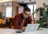 Contemplating businessman in the home office feeling frustrated. Full concentration on work. Man working on laptop, counting profit while sitting at the desk at home.